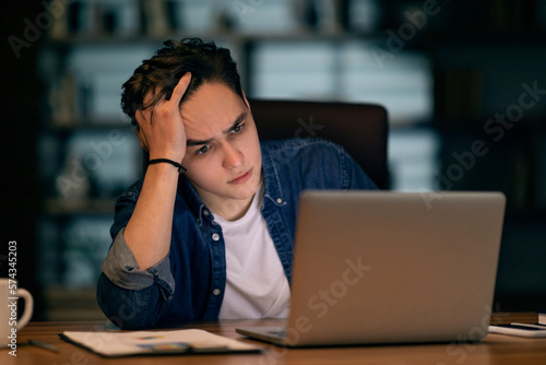 Hopeless frustrated man looking at laptop screen, dark office interior