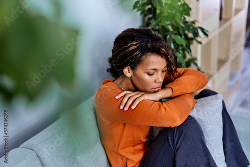 A pensive African woman looking down, sitting on the couch, feeling depressed.