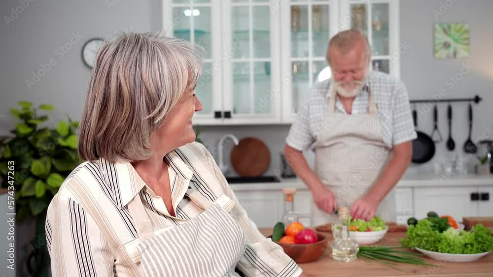 years of marriage, portrait of joyful old woman in an apron with glass of wine background of her husband cutting salad for romantic dinner in kitchen, smiling and looking at camera years of marriage, portrait of joyful old woman in an apron with glass of wine background of her husband cutting salad for romantic dinner in kitchen, smiling and looking at camera