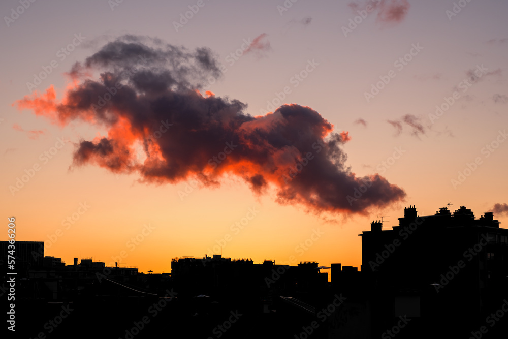 A cloud lit up at sunset passing over the silhouetted chimney stacks and rooftops of Paris in France