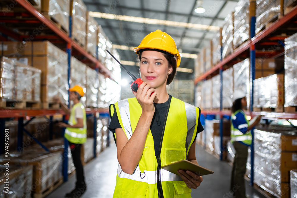 Happy Professional woman Worker Wearing Safety Vest and Hard Hat Smiling on Camera. In the Background Big Warehouse with Shelves full of Delivery Goods. Happy Professional woman Worker Wearing Safety Vest and Hard Hat Smiling on Camera. In the Background Big Warehouse with Shelves full of Delivery Goods.