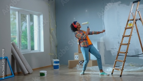 Young african american woman in big wireless red headphones is enjoying music. Black female in checkered shirt sings into paint roller as if into microphone and dances. Ladder, cardboard boxes, window