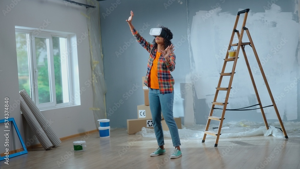 African american woman in virtual reality headset furnishes interior of room. Female makes choice of wallpaper, furniture, curtains using virtual technology. Concept of modern apartment renovation.