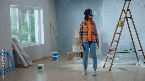 Young african american woman in virtual reality headset visualizing interior of room. Female in checkered shirt makes choice using virtual technology. Concept of modern apartment renovation.