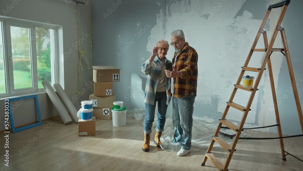 Elderly man and woman are talking on a video call using their mobile phone. Aged couple communicates with someone and showing repairs in the apartment, ladder, cardboard boxes, a window and wallpaper. Elderly man and woman are talking on a video call using their mobile phone. Aged couple communicates with someone and showing repairs in the apartment, ladder, cardboard boxes, a window and wallpaper.