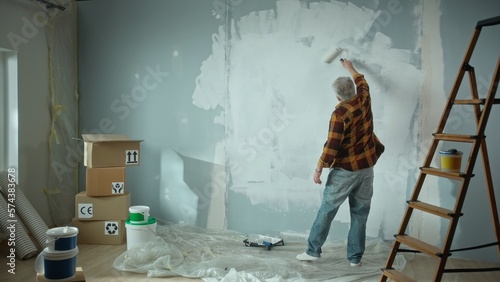 Elderly man is painting wall with white paint using paint roller. Pensioner is making repairs to their apartment, in the background of a window, stepladder, cardboard boxes, wallpaper and paint. Back