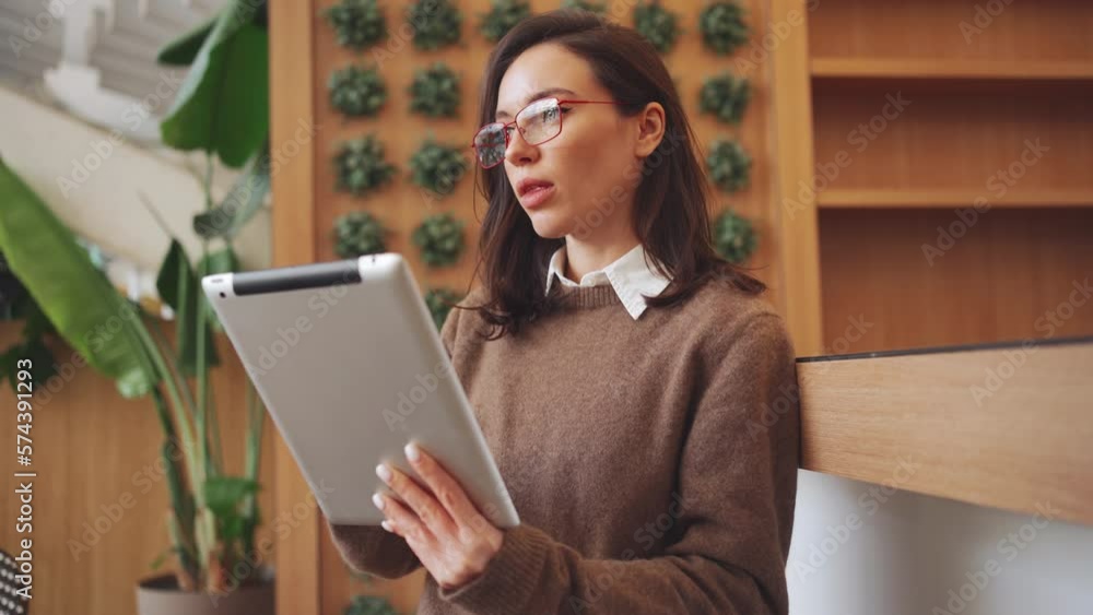 Businesswoman using digital tablet in office at work. Serious female entrepreneur wearing glasses standing and using wireless technology startup business calling someone by finger gesture. Side view