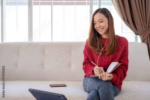 Bright and cute Asian woman sitting on sofa with laptop to work and happily taking notes at home.