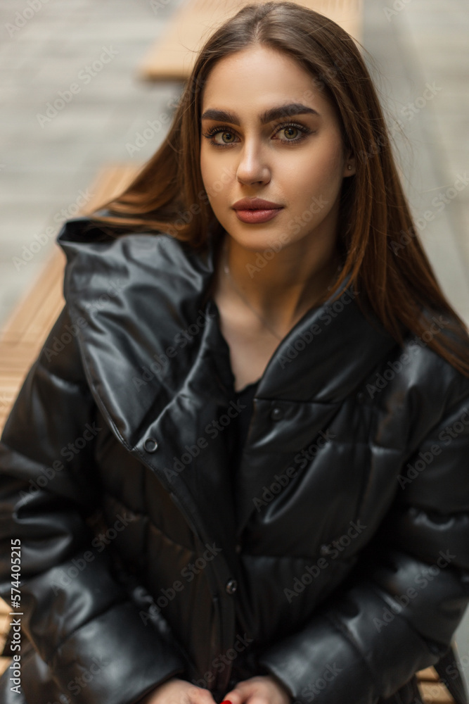 Beautiful young fashion woman in a stylish black winter down jacket sits on a bench and looks at the camera