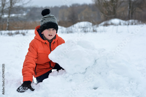 A boy in a jacket and a hat sits in the snow and looks into the distance with surprise. Winter vacation.