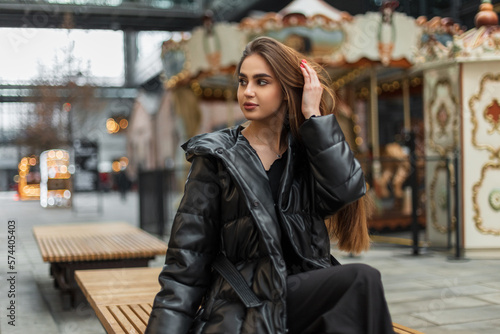 Beautiful young caucasian woman model in a fashion black down jacket with trousers sits on a bench in the city near a vintage carousel
