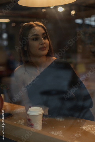 Beautiful young brunette woman in a black top is sitting in a cafe near the window  drinking coffee and waiting for a friend