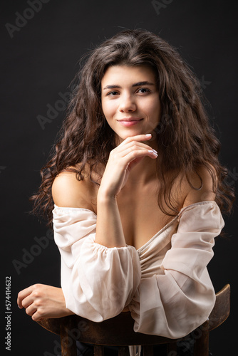 Classic studio portrait of a young brunette in a white loose top, who is sitting on a chair against a black background.