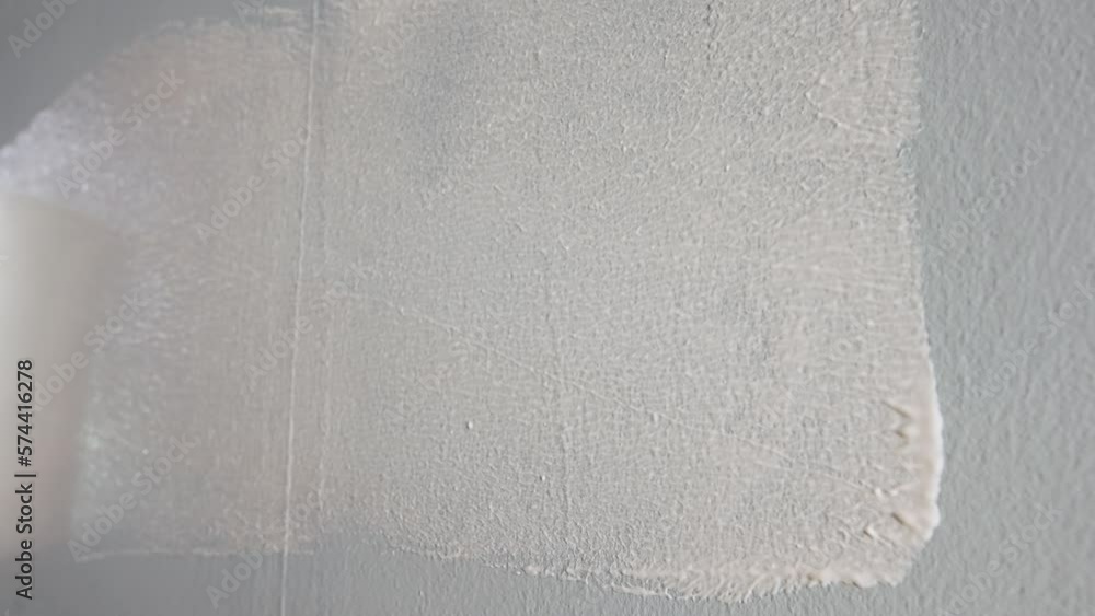 The unrecognizable person paints a gray wall with white paint using a paint roller. A close up of a round, blue handled roller slides over a wall and paints over it. The concept of repair. The unrecognizable person paints a gray wall with white paint using a paint roller. A close up of a round, blue handled roller slides over a wall and paints over it. The concept of repair.