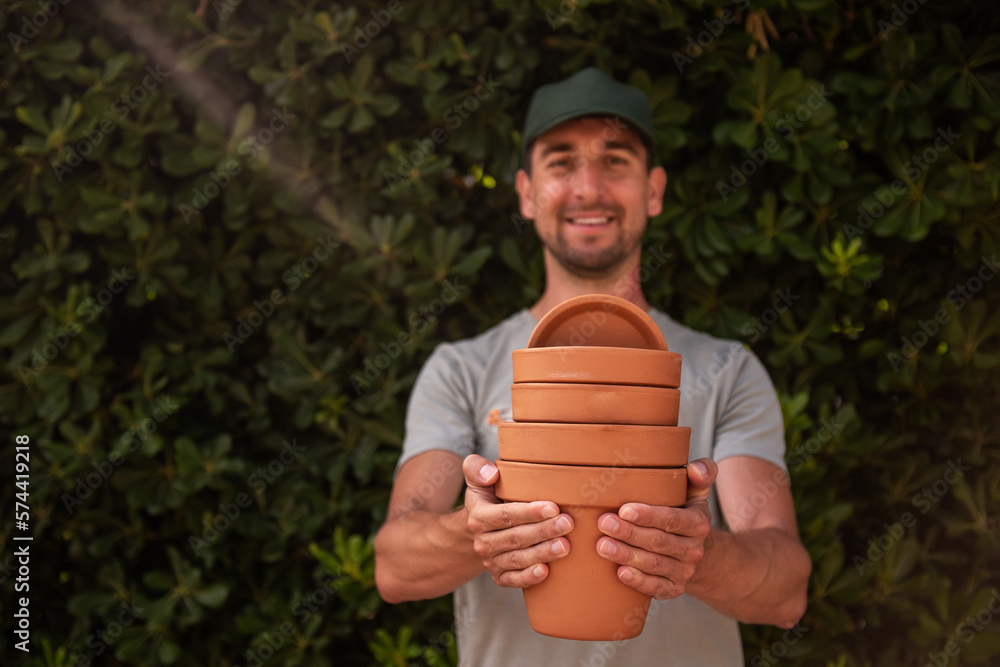 Close-up portrait man gardener in green cap holds terracotta clay pots in hands against the backdrop of an evergreen fence. Gardening, small business, hobby. Trendy garden landscaping. Copy space Close-up portrait man gardener in green cap holds terracotta clay pots in hands against the backdrop of an evergreen fence. Gardening, small business, hobby. Trendy garden landscaping. Copy space