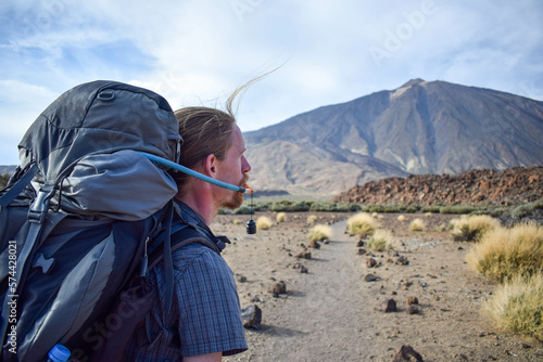 Handsome young traveler man hiking drinking water on mountain with Teide volcano on the background. Enjoying hot weather and sun in Spain. Staying hydrated on sport hike. Hydration bladder.