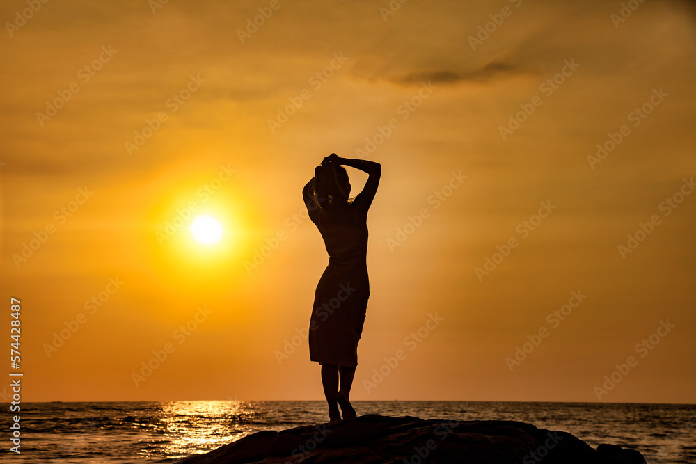 Silhouette young lady in dress standing on rock at tropical ocean background, tropic sunset. Silhouette slim woman enjoy rest on seacoast. Travel vacation holiday concept. Copy advertising text space