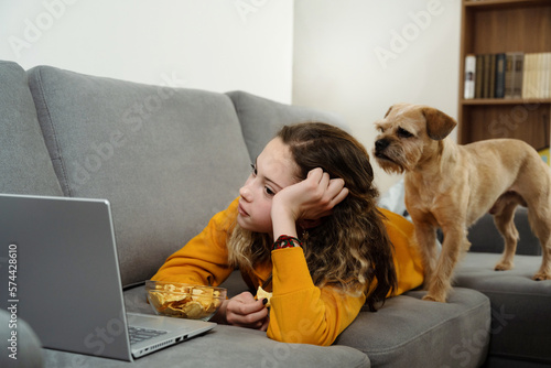 Favorite hobby. Teen girl is watching a movie on a laptop and is lying on the couch at home with dog pet. © Oksana Klymenko