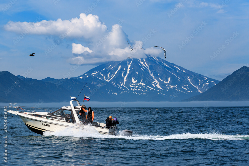 Fototapeta premium A boat with tourists floats in the Pacific Ocean against the backdrop of a snow-covered volcano and mountains. Kamchatka.
