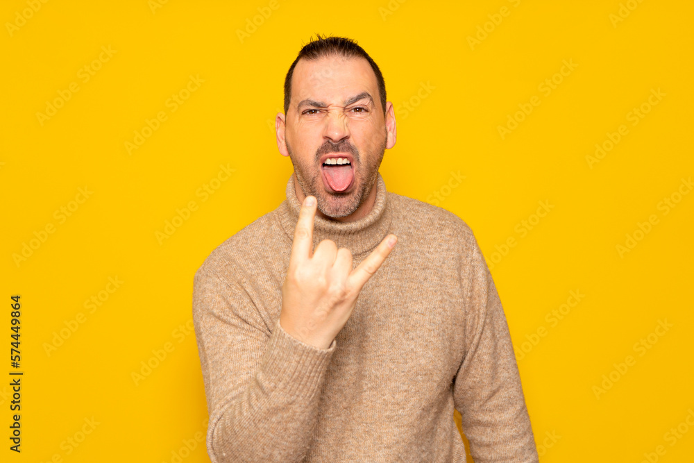 Bearded hispanic man wearing turtleneck isolated over yellow background showing rock gesture with fingers and sticking out tongue. Concept of rebellion and anarchism.