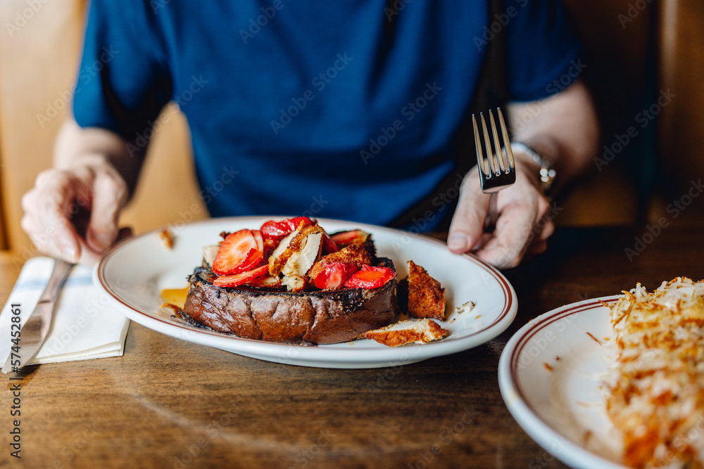 Delicious plate of food with French toast, chicken and strawberries, with man sitting behind plate, holding knife and fork 