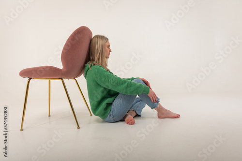 portrait of a young emotional girl sitting on the floor near a chair in a flirtatious mood, gesturing with her hands, empty space, wearing jeans and a green sweater n, space for text