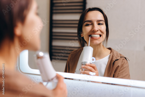 A young girl stands in front of a mirror in the bathroom and rinses her mouth with an irrigator.