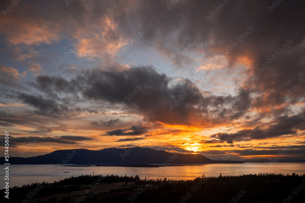 Dramatic Sunset Over Orcas Island, Washington. Aerial view of a Tugboat towing a barge through Rosario Strait between Lummi Island and Orcas Island in the San Juan Islands. Dramatic Sunset Over Orcas Island, Washington. Aerial view of a Tugboat towing a barge through Rosario Strait between Lummi Island and Orcas Island in the San Juan Islands.