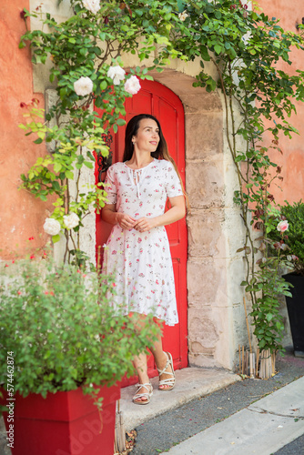 Lifestyle portrait of young stylish woman staying near red door on the street in old town and looking at white roses flowers, wearing white midi dress © svittlana