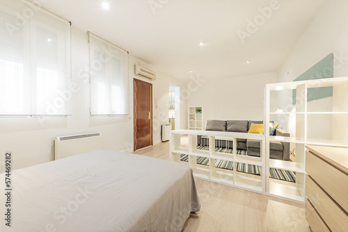 Loft-style apartment furnished with white wooden shelves, chest of drawers, armored access door and wooden floors covered with jarapa carpet and double bed with gray bedspread photo