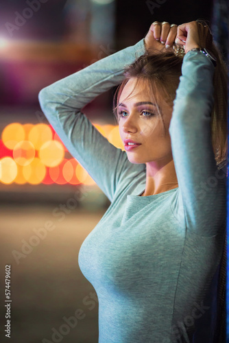 Close-up portrait of a beautiful slim blonde caucasian girl looking left sexually posing, shot in amusement park with colored bulb lights at the background blurred in huge bokeh and lens flares