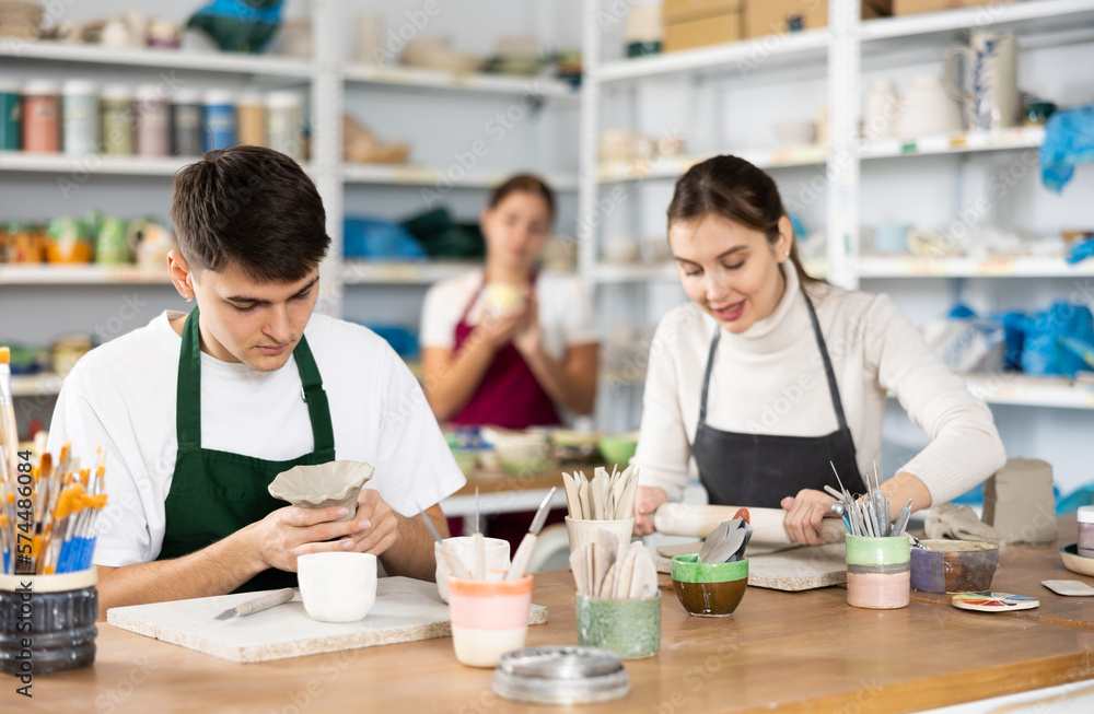 Fototapeta premium Cheerful young man and woman in aprons modeling handmade mugs of row clay material in pottery workroom