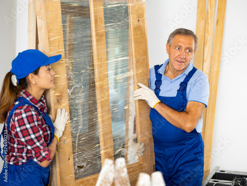 Latin woman and caucasian middle-aged man repairers carrying wooden door together during renovation works in apartment.