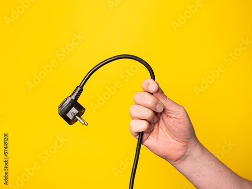 A hand holds a plug to which a black cable is attached. No face, yellow background. 