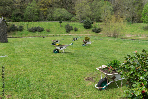 wheelbarrow-shaped pots full of plants and flowers of different colors resting on a ground full of natural green grass, near the Rato river in Lugo. Galicia. Spain
