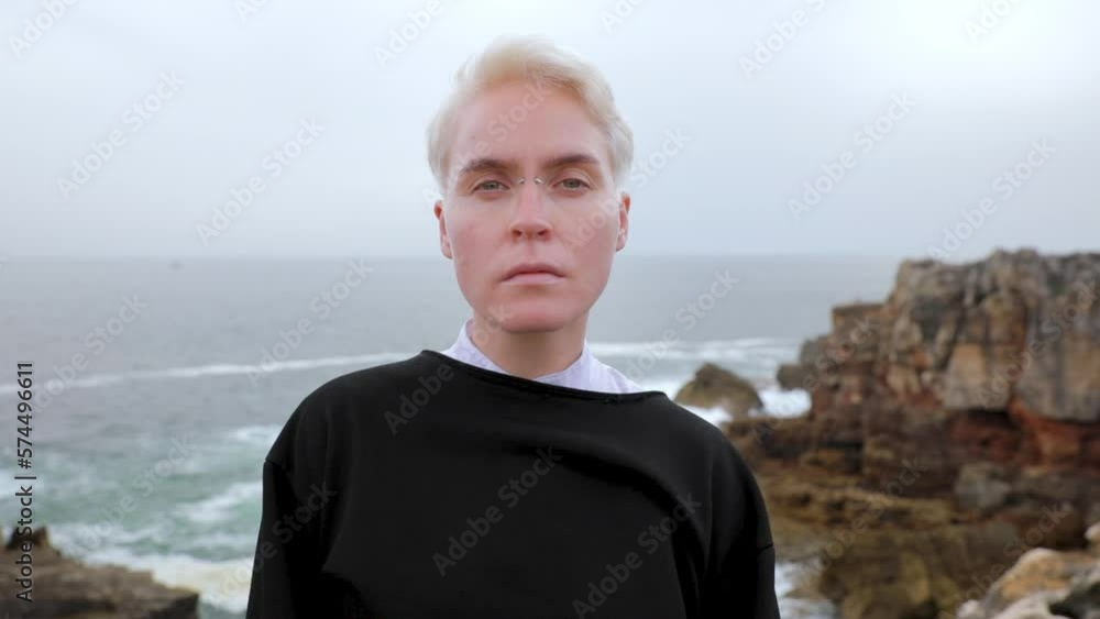 Horizontal video. Close-up portrait of a white girl with short white hair against the backdrop of rocks and the ocean. Looks into the frame and around. Horizontal video. Close-up portrait of a white girl with short white hair against the backdrop of rocks and the ocean. Looks into the frame and around.