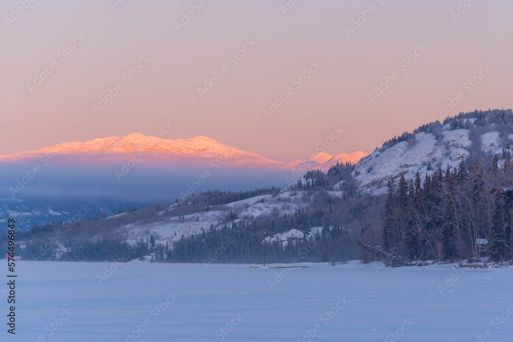 Landscape winter views in northern Canada during sunrise with pink tones on surrounding mountains and sky. Frozen lake in foreground. 