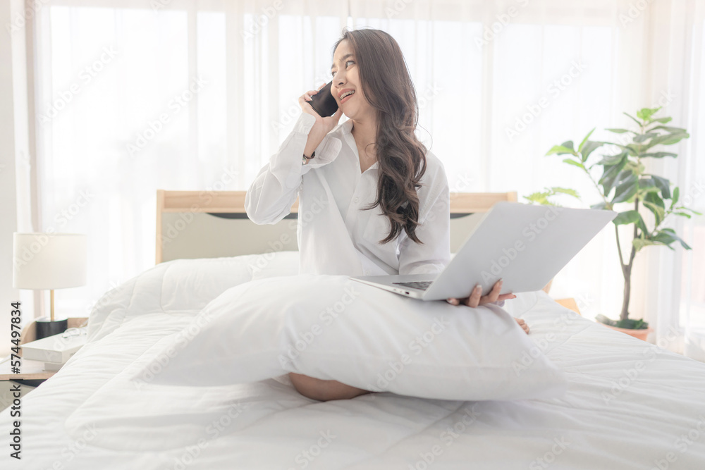 young asian woman using smartphone and laptop when sitting on a bed ...