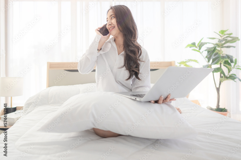 young asian woman using smartphone and laptop when sitting on a bed ...