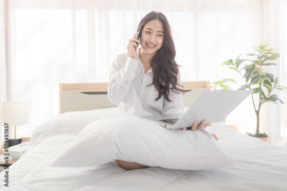 young asian woman using smartphone and laptop when sitting on a bed ...