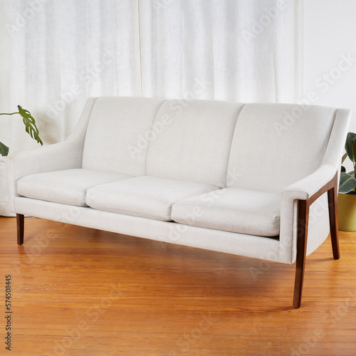 Vintage white sofa. Stylish mid-century modern couch with sculptural wooden legs. Interior scene with white curtains and wooden floor.