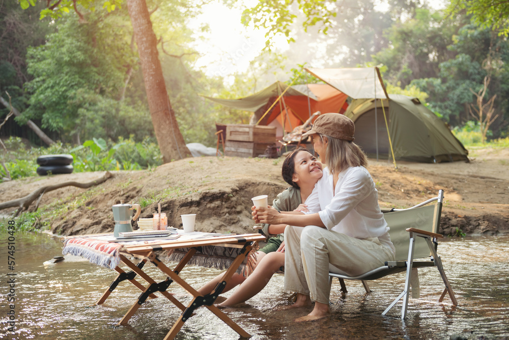 Pleased happy mother and daughter reading a book and using laptop while relaxing on the deck chairs in the river, sit near a camp and tent, drink coffee in a pine forest. Camping, recreation, hiking. Pleased happy mother and daughter reading a book and using laptop while relaxing on the deck chairs in the river, sit near a camp and tent, drink coffee in a pine forest. Camping, recreation, hiking.