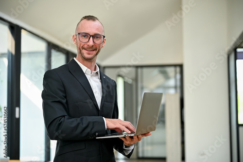Confident Caucasian businessman looking at camera while standing in the office with his laptop.