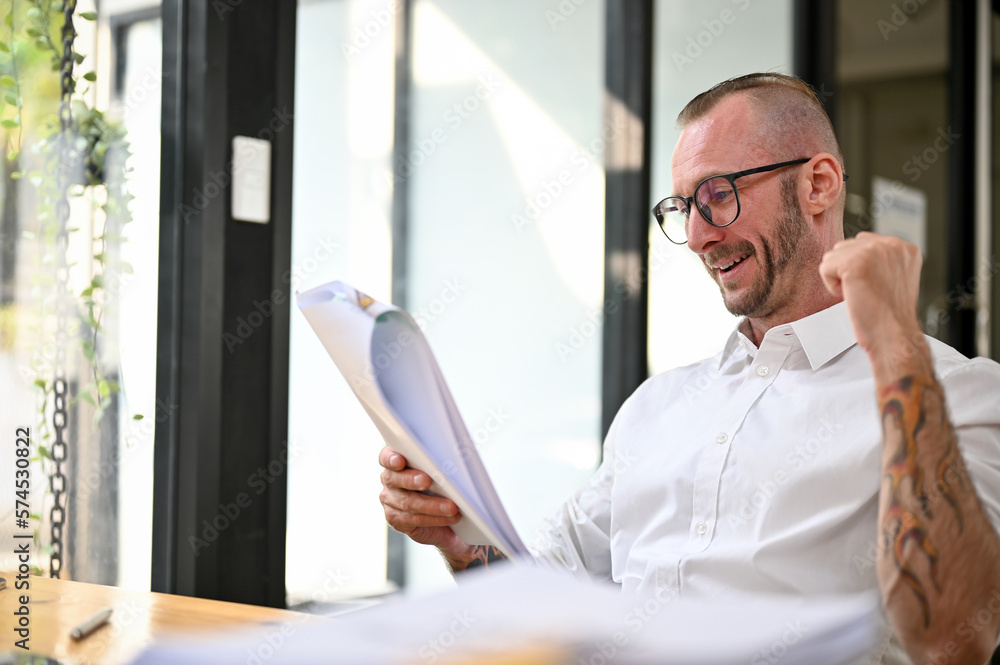 Fototapeta premium Happy Caucasian businessman looking at business paperwork, showing yes gesture. good news