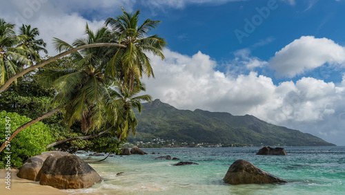 Beautiful tropical beach. Granite boulders in the turquoise ocean and on the shore. Coconut palms bent over the water. A green hill against a background of blue sky and clouds. Seychelles. Mahe