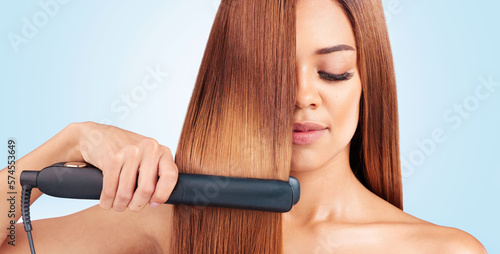 Face, beauty and woman with hair straightener in studio isolated on a blue background. Haircare, wellness and young female model with flat iron for salon treatment hairstyle, aesthetic or balayage. photo