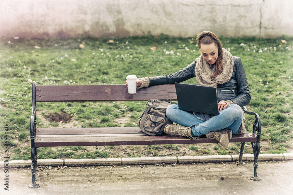 Long hair hipster brunette woman sitting on a bench in the park,  working on a laptop and drinking a cup of coffee.