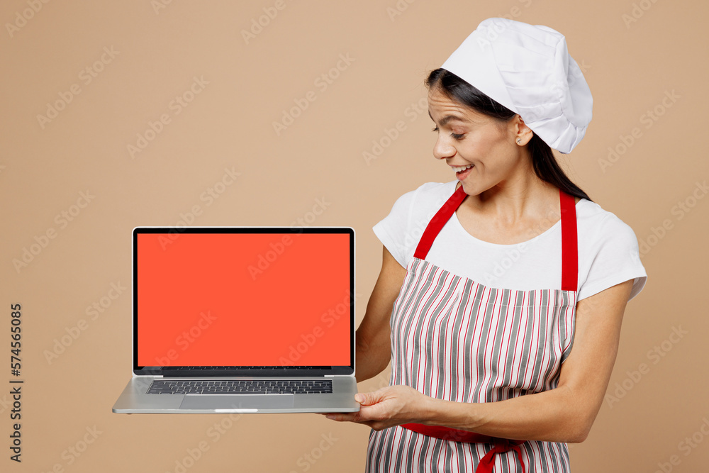Young housewife housekeeper chef baker latin woman wear striped apron toque hat work hold use black screen laptop pc computer look for recipe isolated on plain light beige background Cook food concept Young housewife housekeeper chef baker latin woman wear striped apron toque hat work hold use black screen laptop pc computer look for recipe isolated on plain light beige background Cook food concept