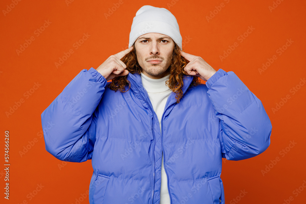 Young sad man with long curly hair wear hat purple ski padded jacket casual clothes cover ears with hands fingers do not want to listen scream isolated on plain orange red background studio portrait. Young sad man with long curly hair wear hat purple ski padded jacket casual clothes cover ears with hands fingers do not want to listen scream isolated on plain orange red background studio portrait.
