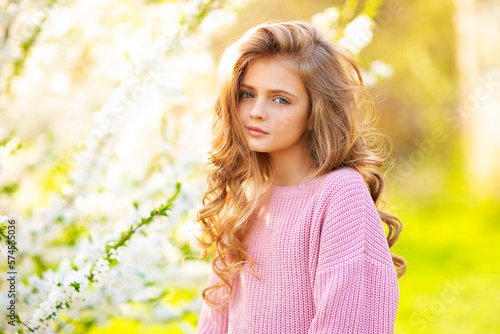 Portrait of beautiful young blonde curly girl with blue eyes posing on spring nature with white blooming trees.Sunny positive colorful photo with amazing teenage model. Light in hair.  